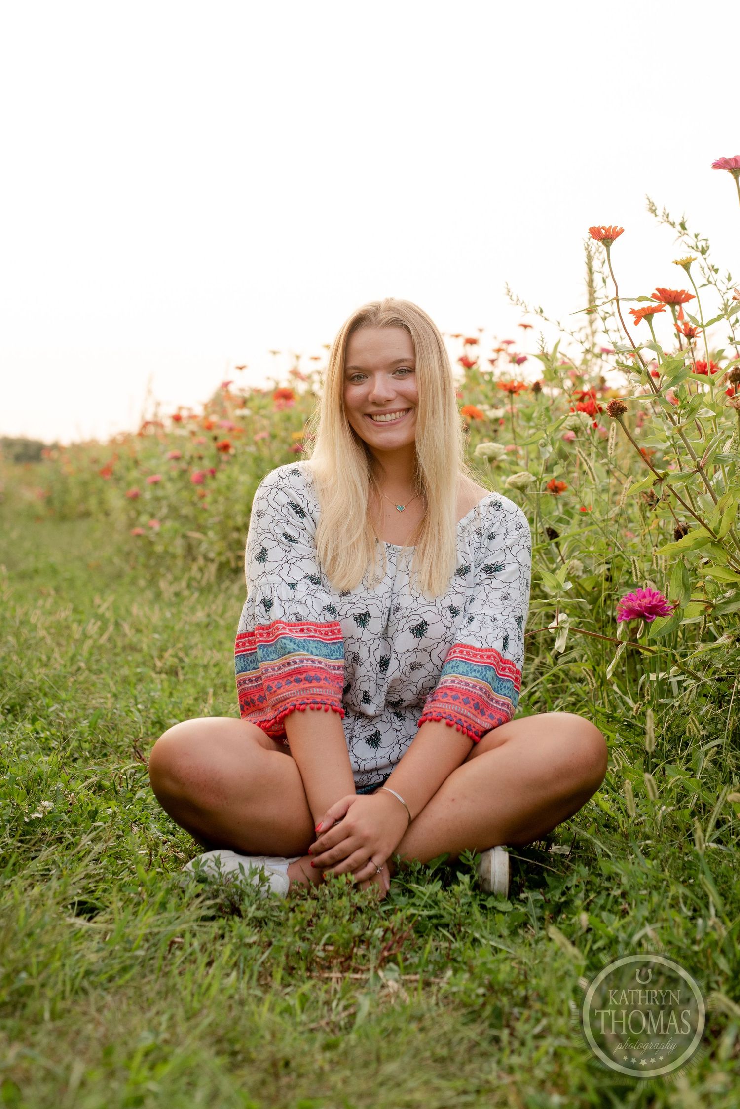 A woman sits cross-legged in a field of red flowers wearing a gray and coral patterned dress.