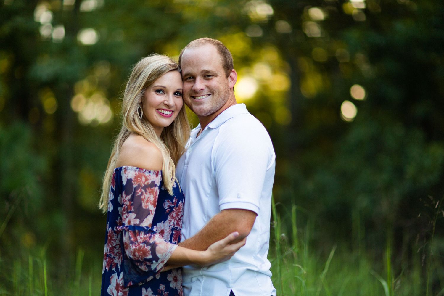 Couple in casual attire poses together against green foliage background.