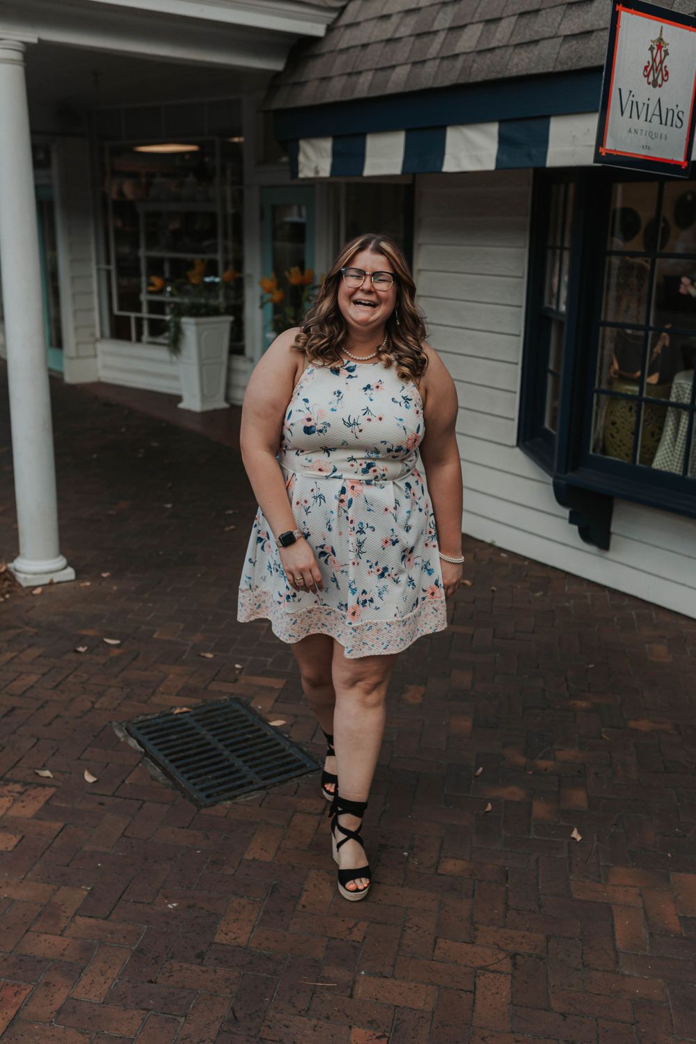 Graduate in floral dress walks cheerfully along brick pathway.