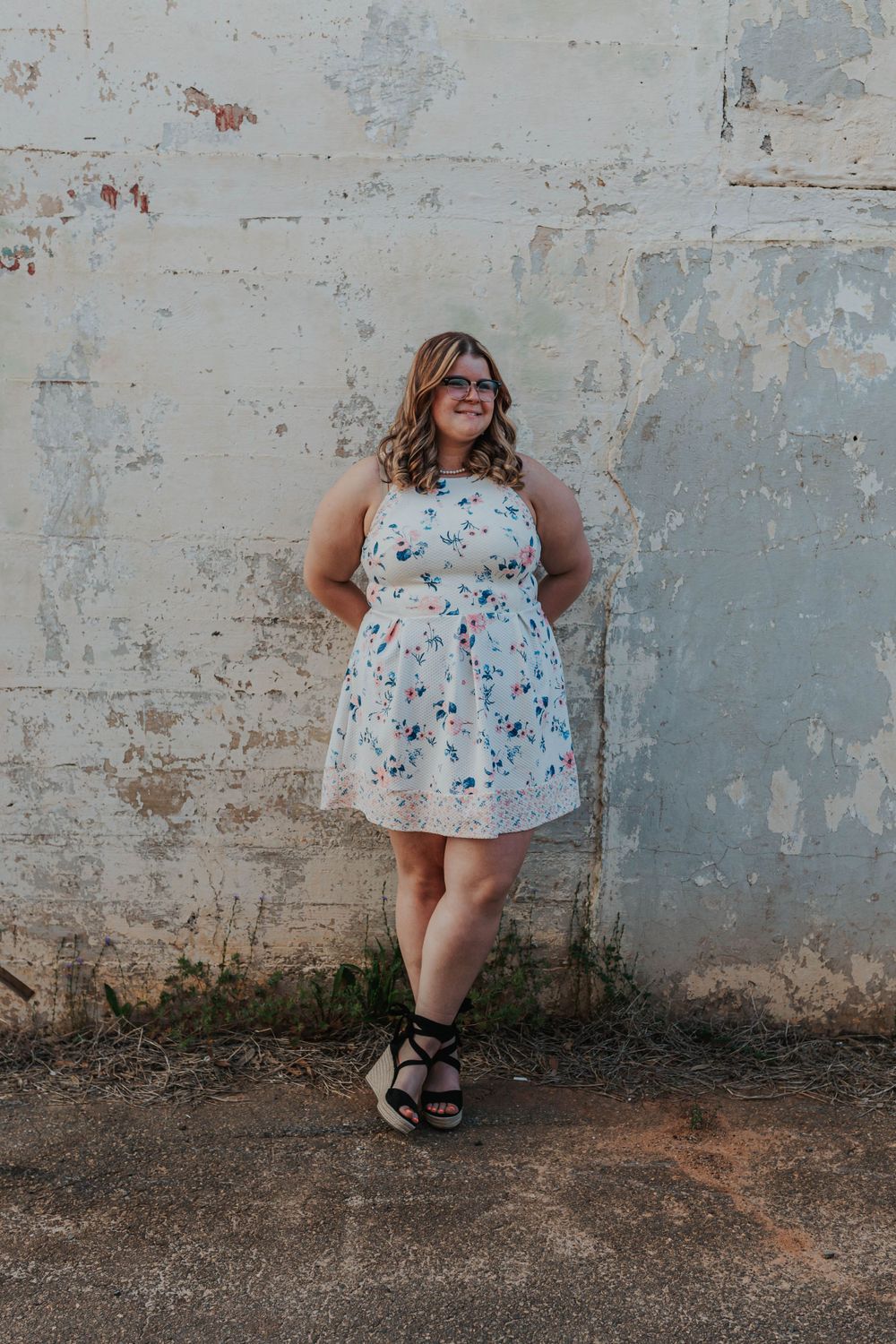 Person in white floral dress poses playfully against distressed concrete wall.