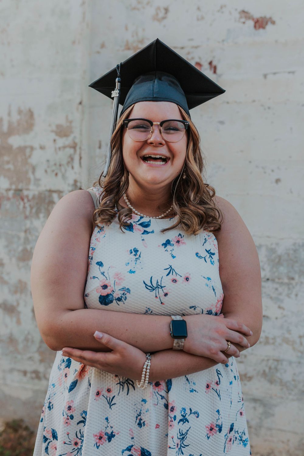 Graduate in floral dress and cap poses joyfully against weathered wall.