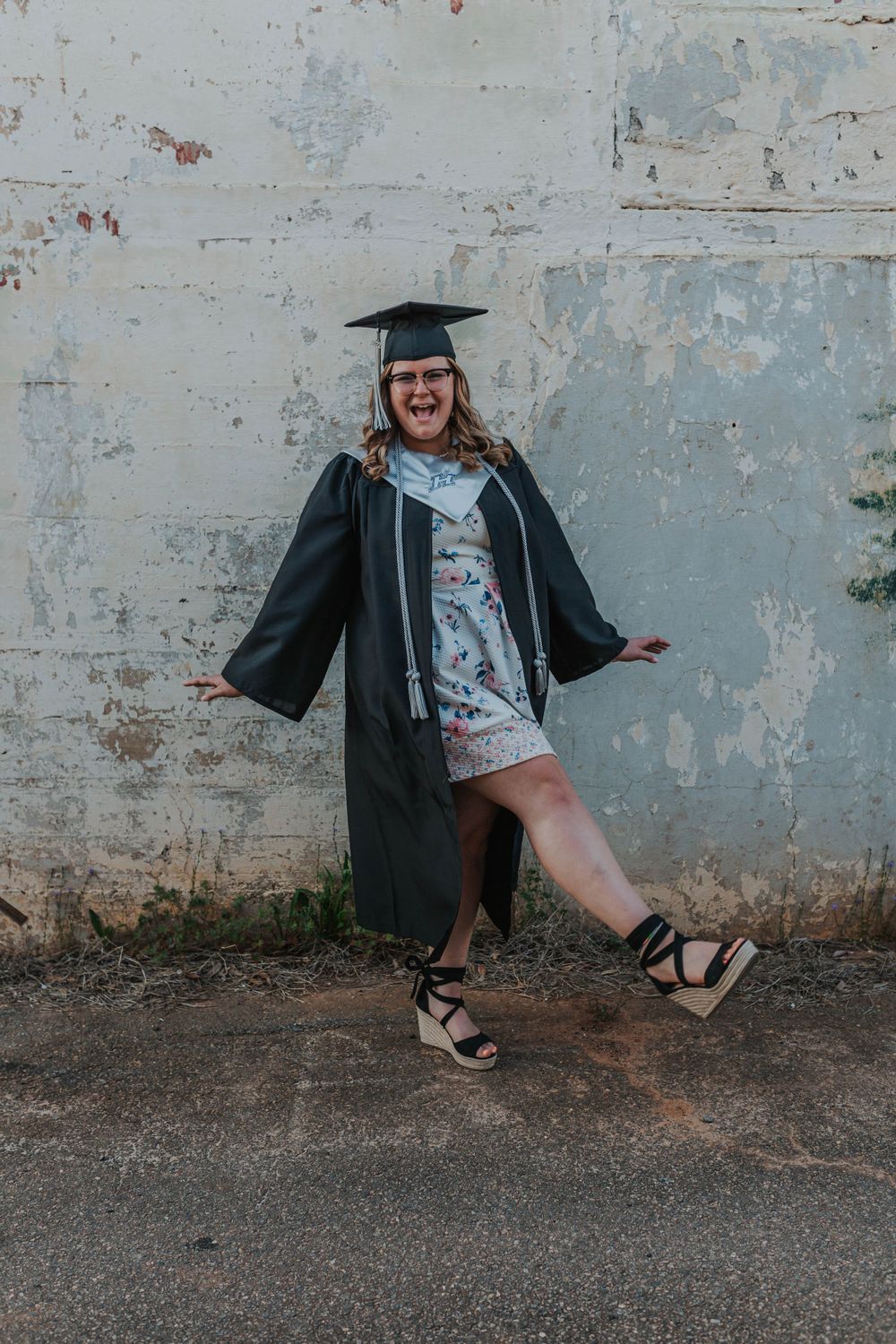Graduate in cap and gown strikes fun poses against concrete wall.