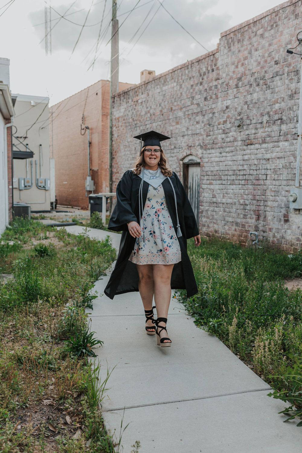 Graduate walks confidently down concrete path near brick buildings.