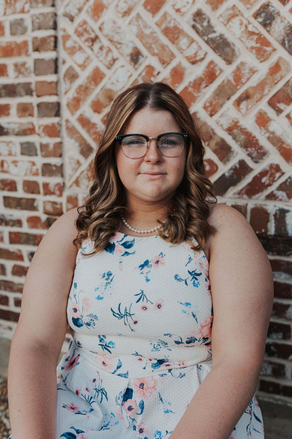 Graduate in floral dress and glasses poses against brick wall background.