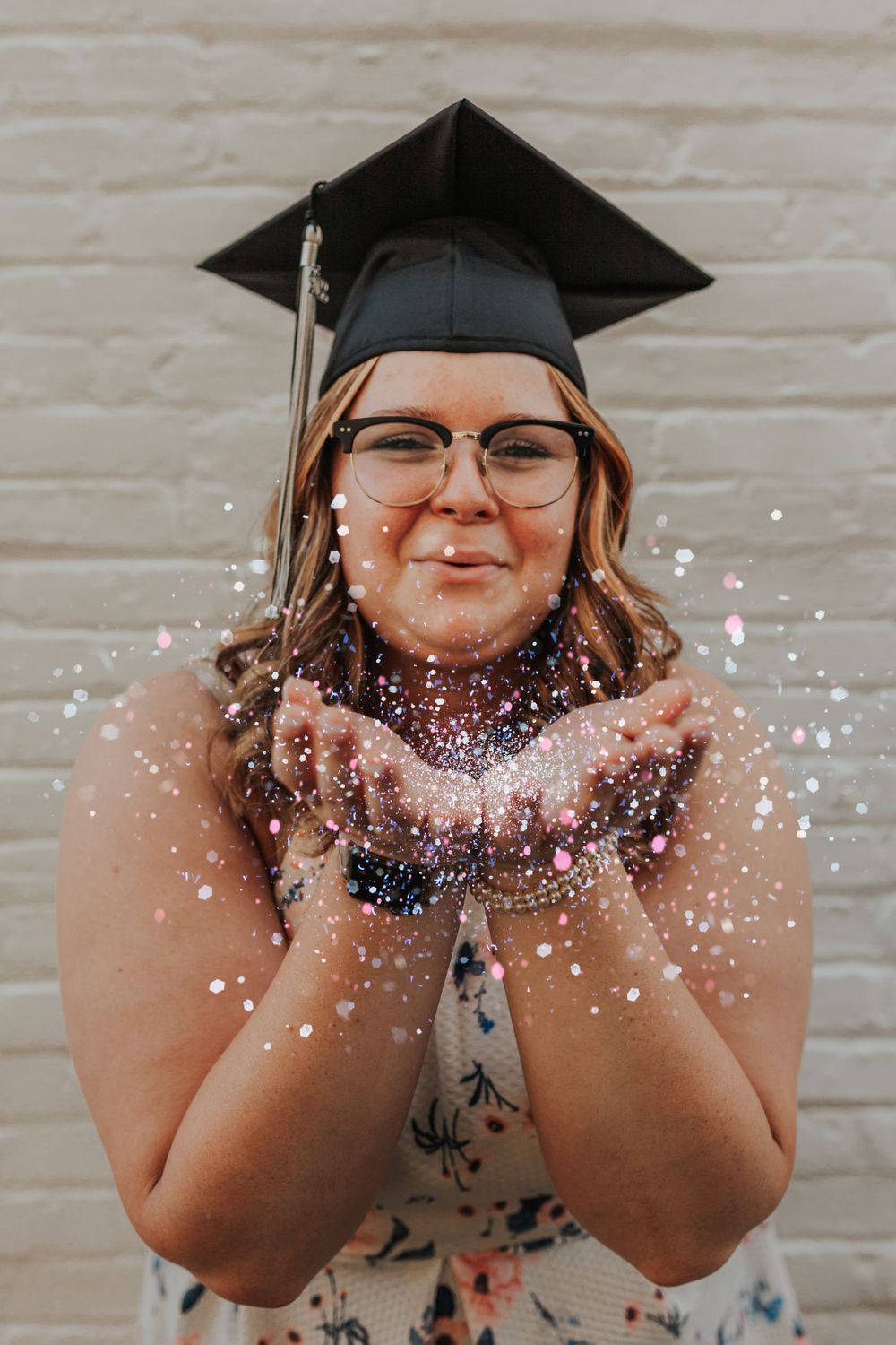 Graduate in cap blows glitter from hands in celebratory pose.