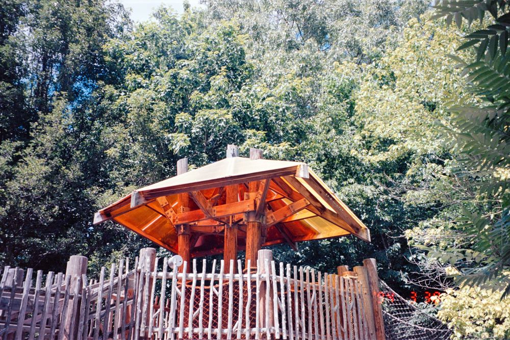 A wooden gazebo with red-painted details sits atop a fenced platform surrounded by trees.