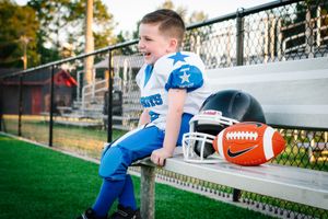 Young football player in blue uniform sits on bleachers next to ball and helmet.