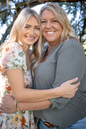Two women embrace and smile warmly while posing outdoors under a tree.