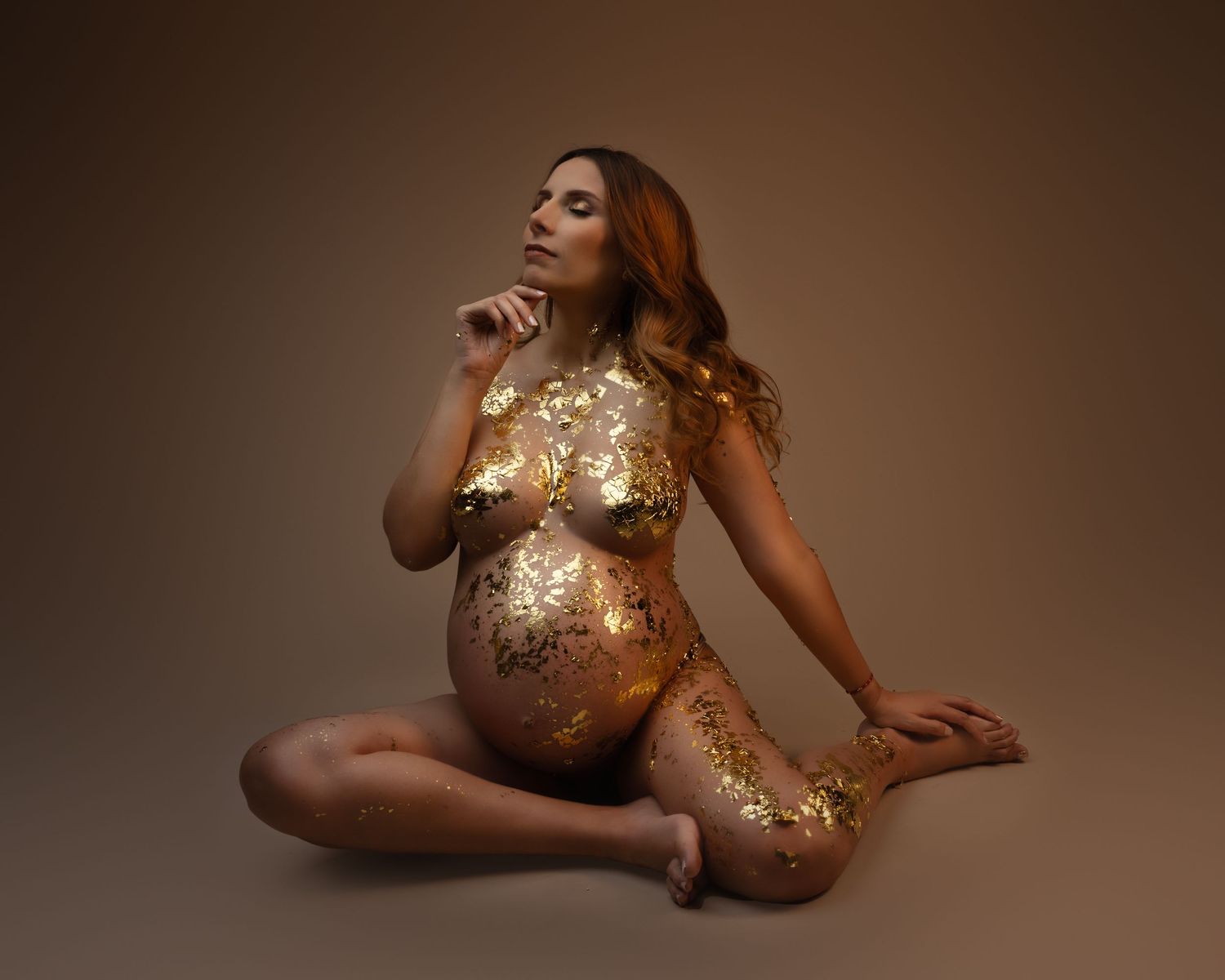Artistic maternity photo featuring metallic gold body paint and glitter in a minimalist studio setting.