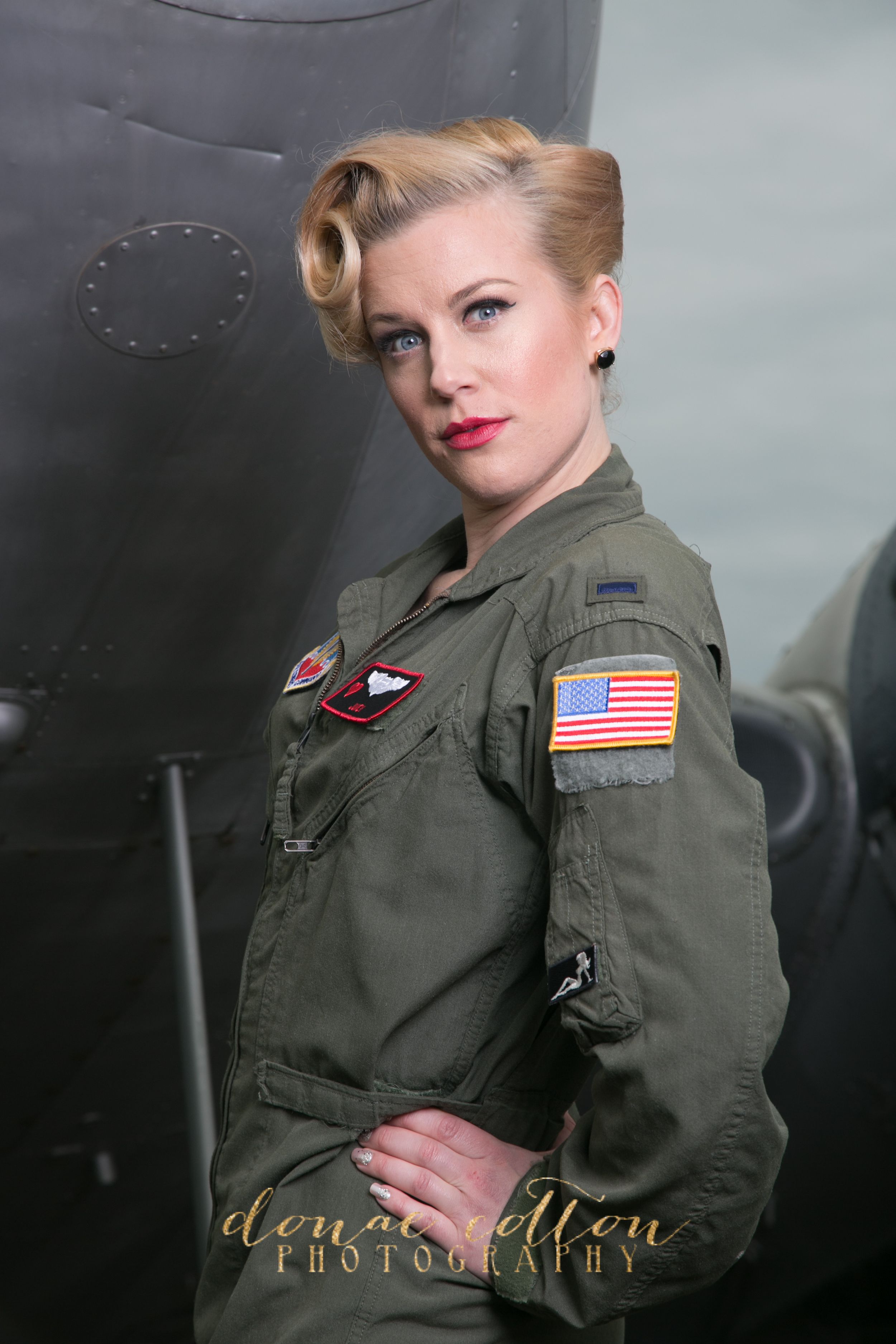 Retro pin-up blonde woman in olive green military flight suit with US flag patch by an aircraft.