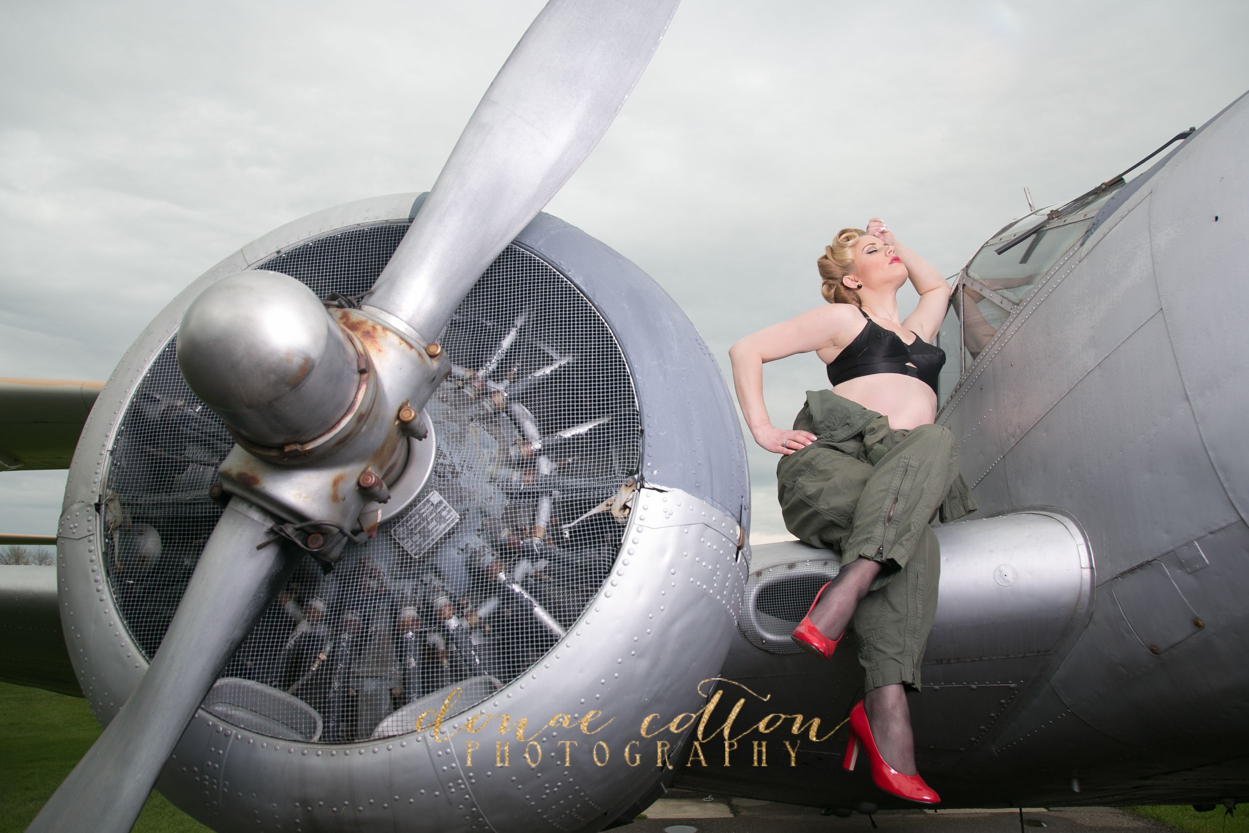 Vintage pin-up woman in black crop top and olive pants poses beside a large aircraft engine.