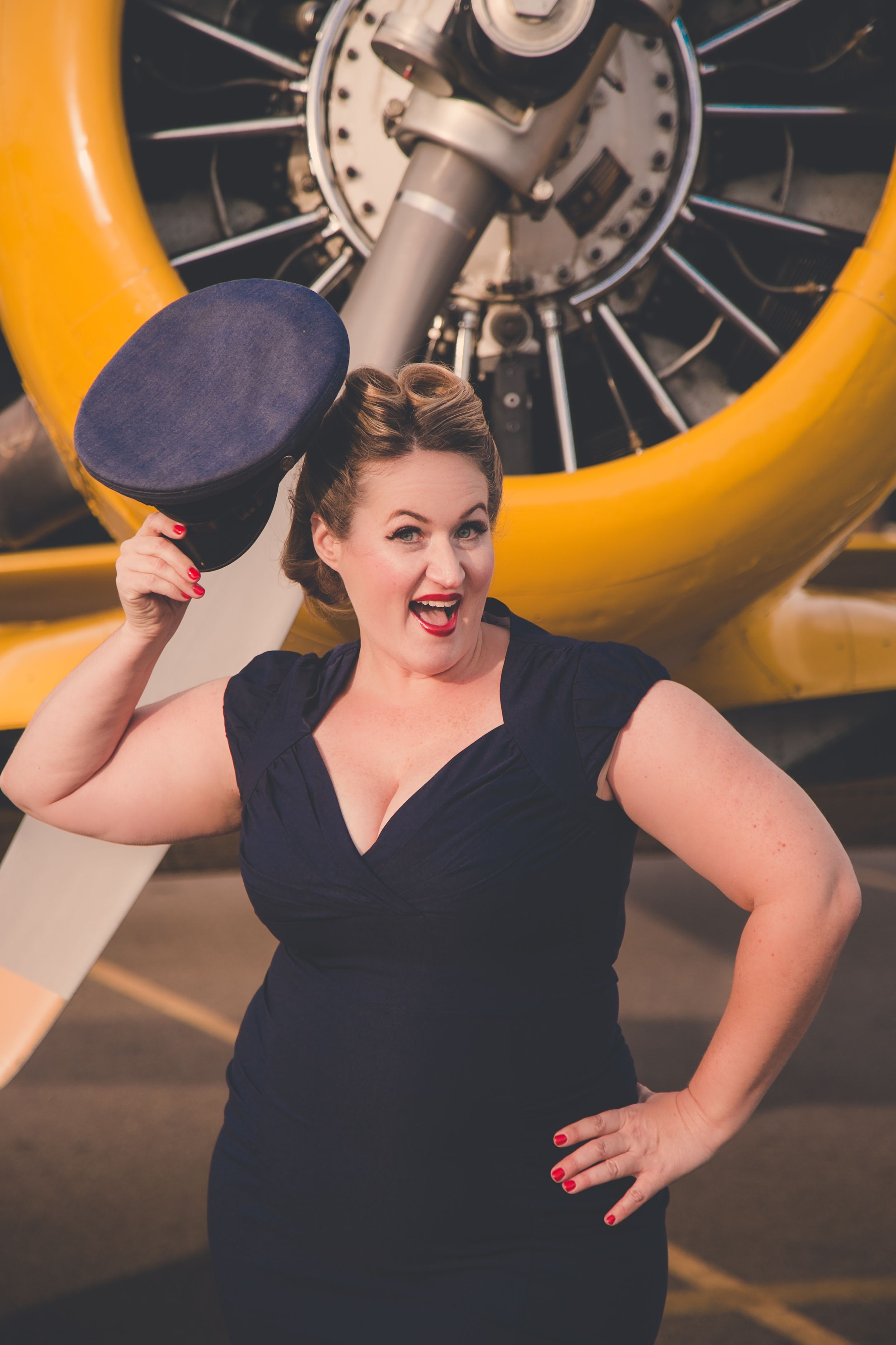 Woman in navy dress holding a cap while posing in front of a yellow vintage aircraft engine.