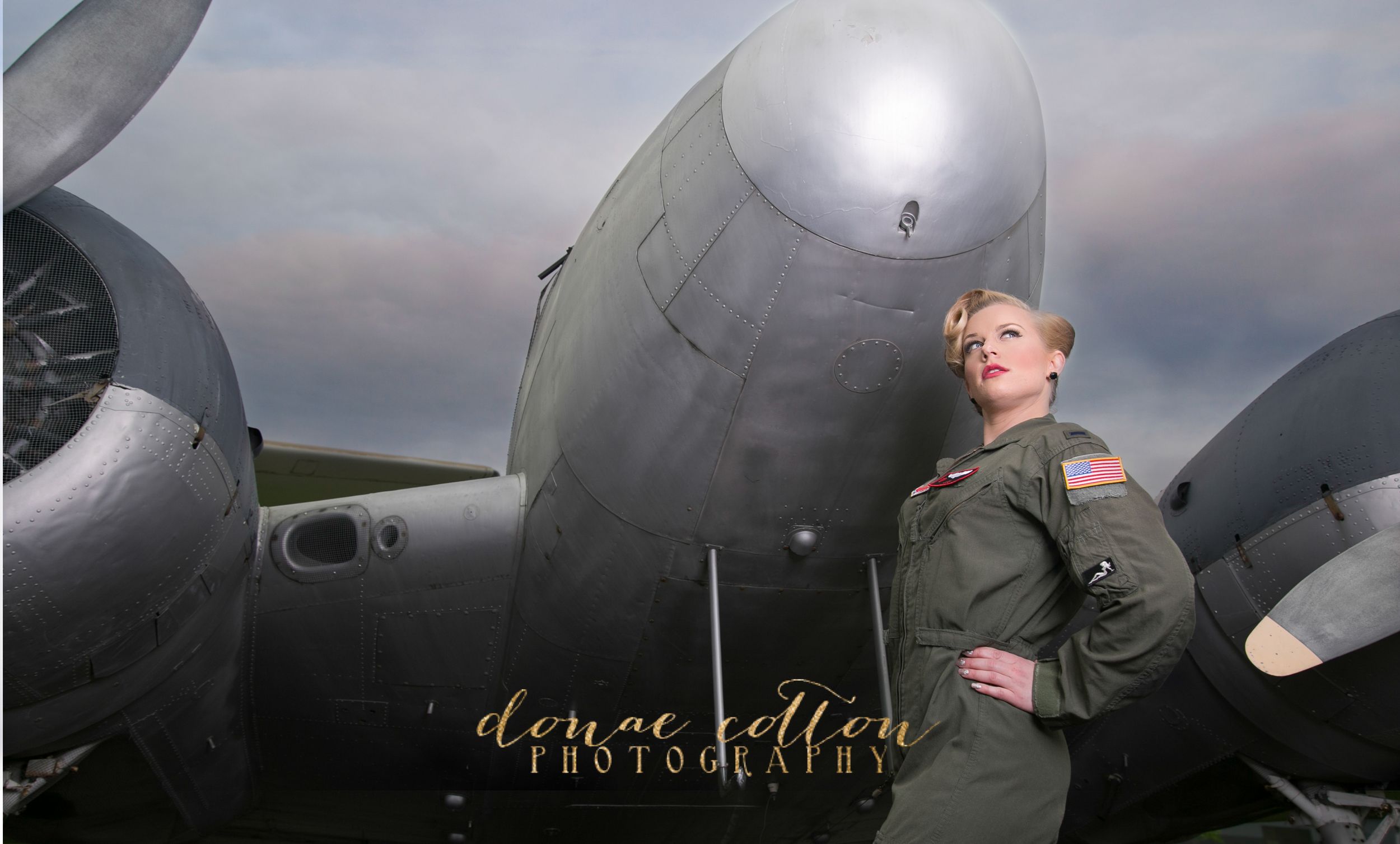 Woman in green flight suit poses beside a large vintage WWII bomber aircraft on a runway.