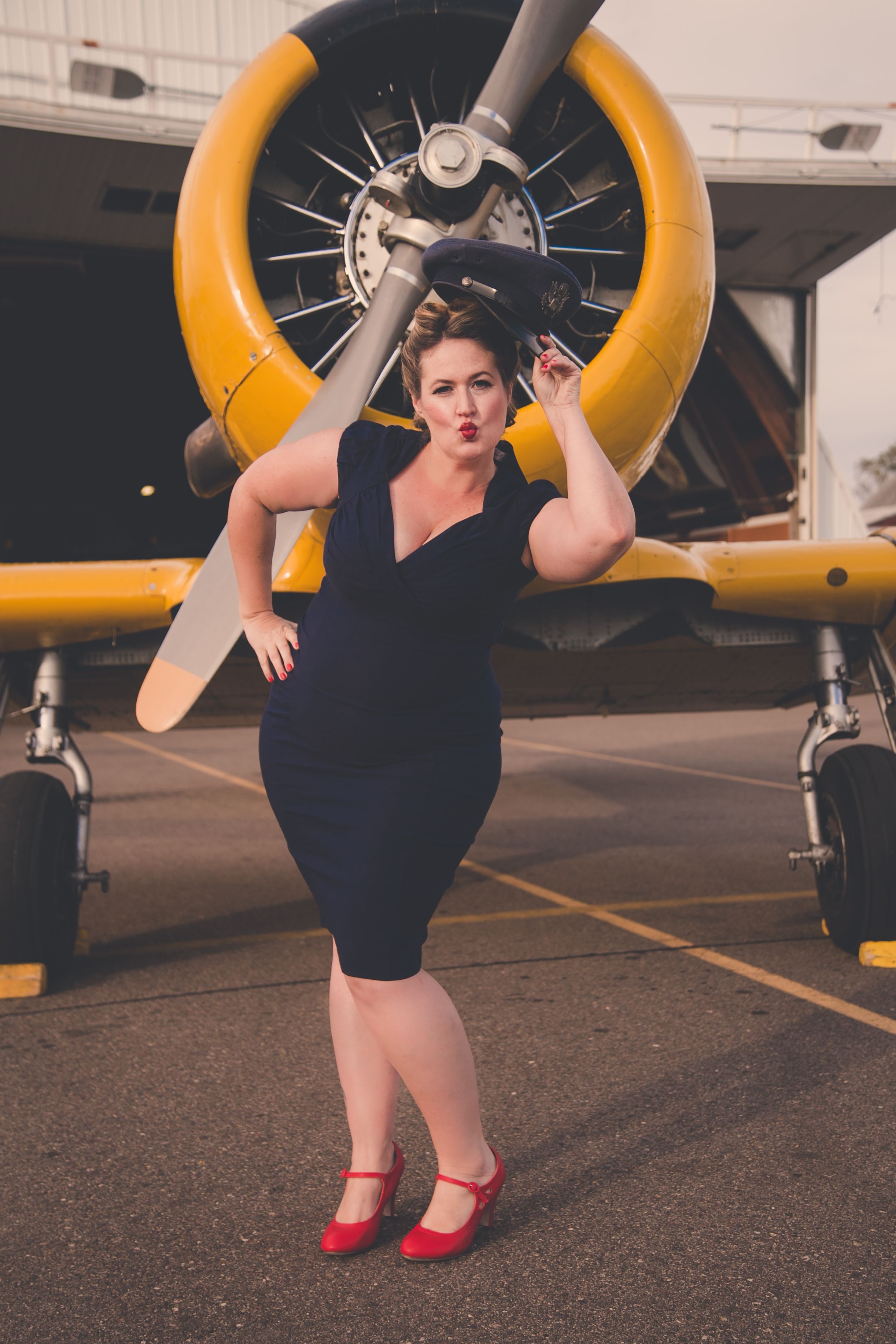 Woman in navy dress posing in front of a yellow vintage military aircraft propeller outdoors.