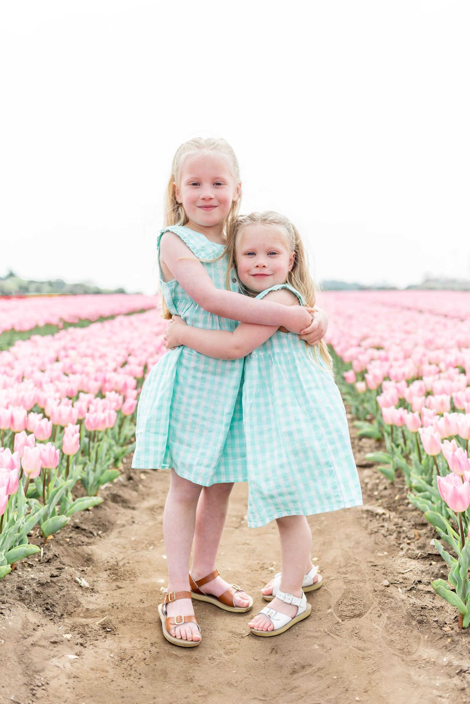 Two young girls in matching mint gingham dresses hugging in a tulip field.