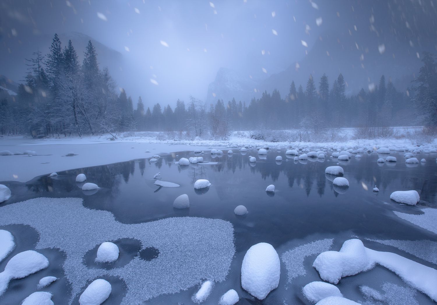 Snow-covered rocks surround a partially frozen lake in a misty winter forest landscape during twilight.