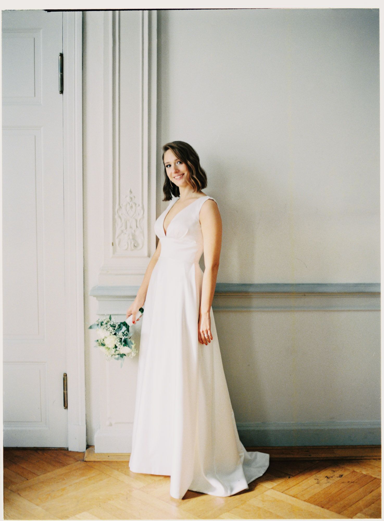 An elegant bride in a classic white wedding gown poses against a light colored wall holding a small bouquet.