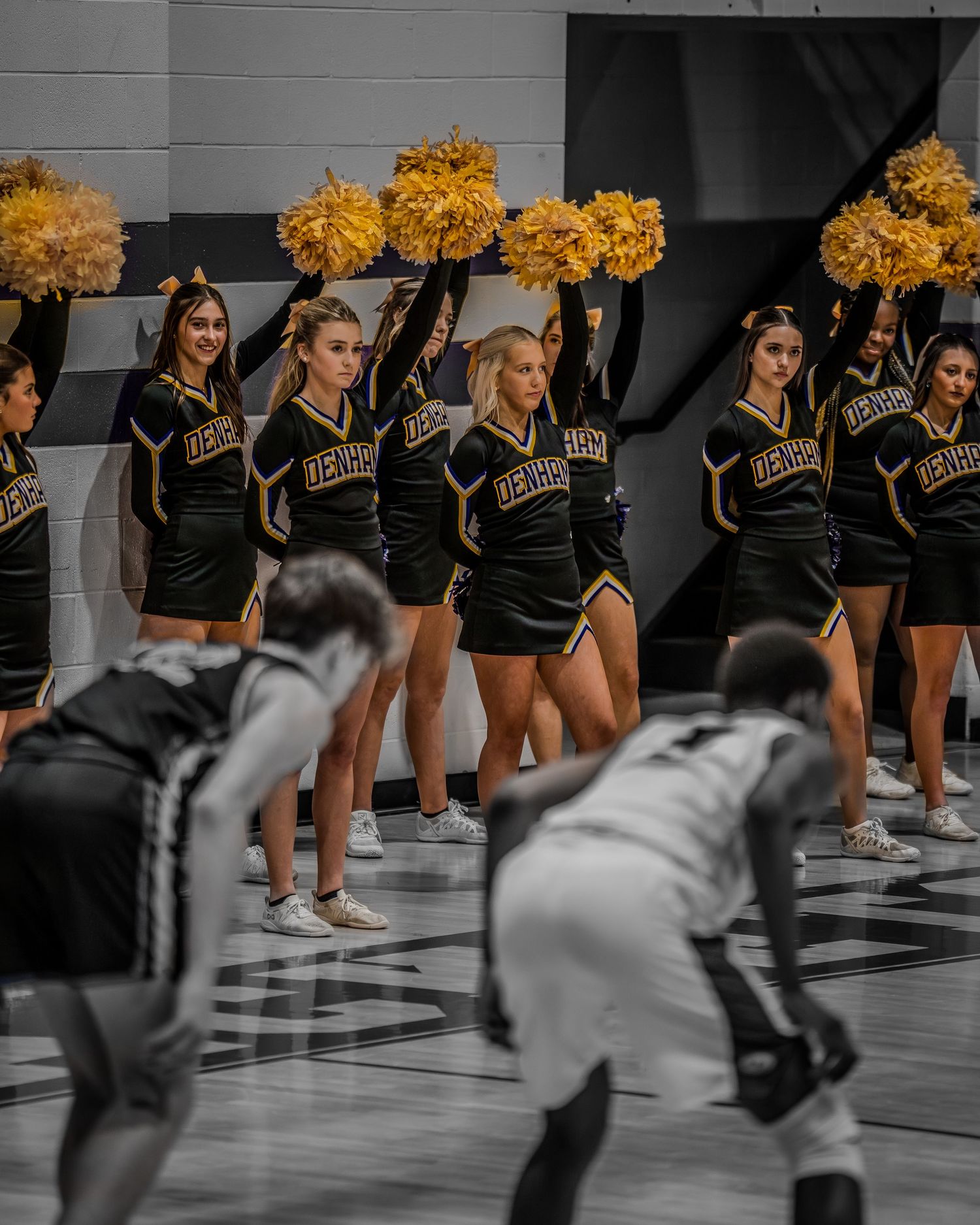 Cheerleaders perform with gold pom poms during a basketball game on a gymnasium court.