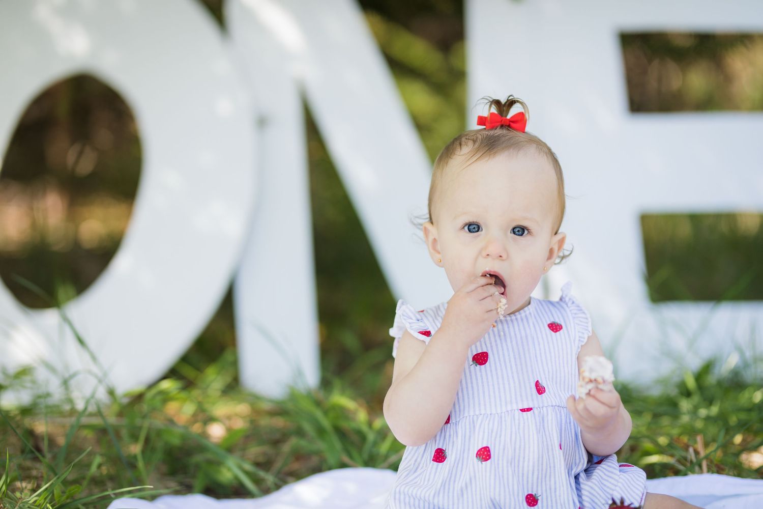 Baby in polka dot dress sits on white blanket outdoors with finger in mouth.
