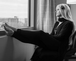 Person in black clothing sitting by window with feet up in black and white photograph.