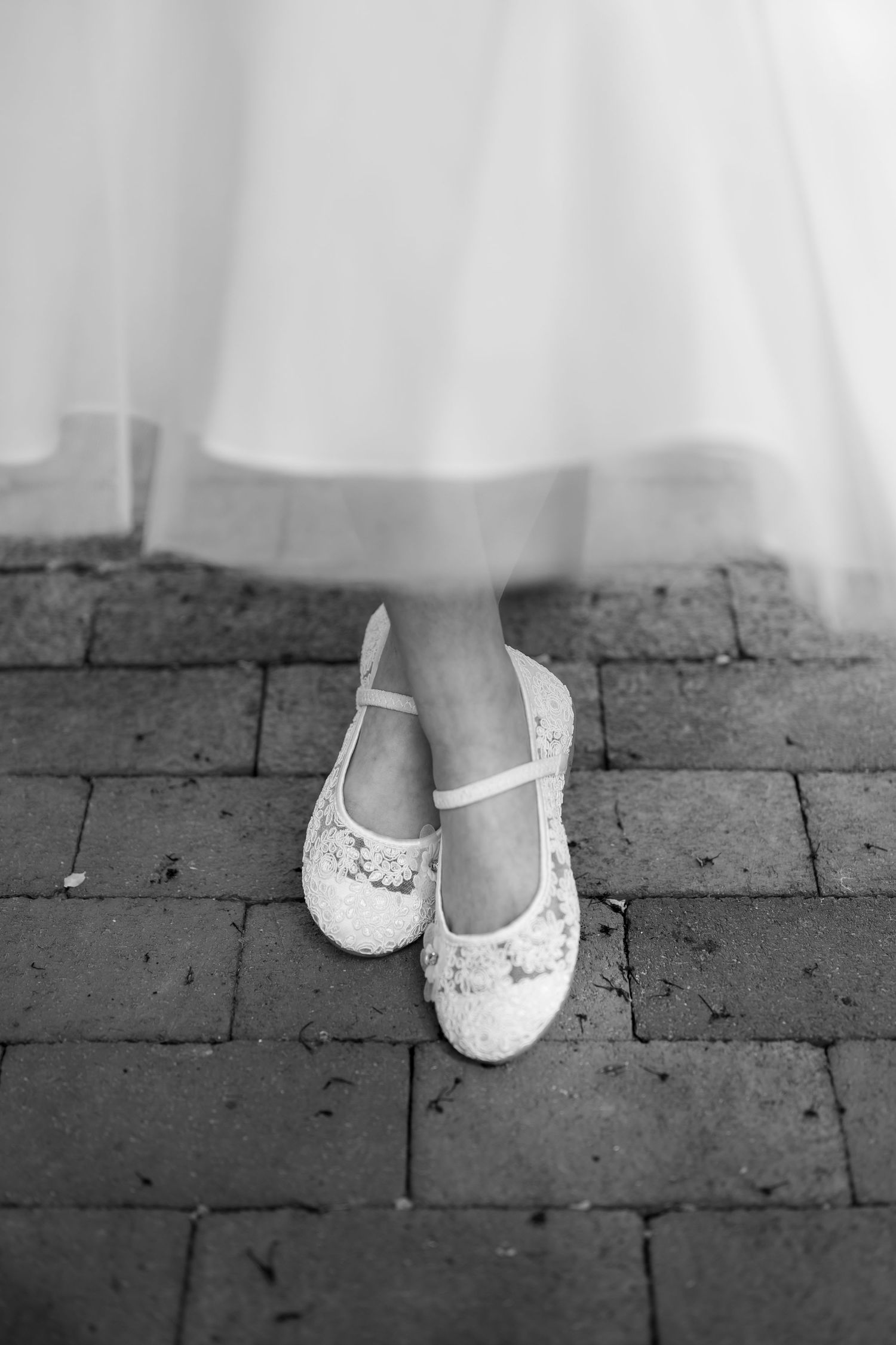 Delicate white lace ballet flats on brick pavement beneath a flowing white dress.