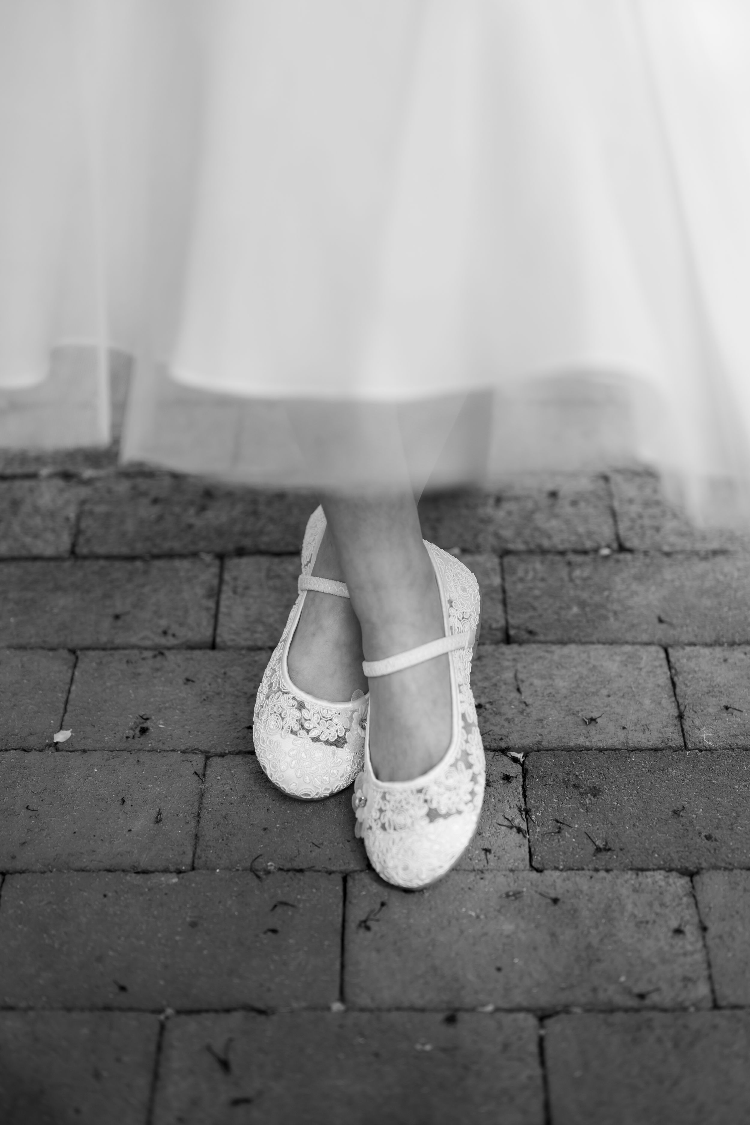 Delicate white lace ballet flats on brick pavement beneath a flowing white dress.