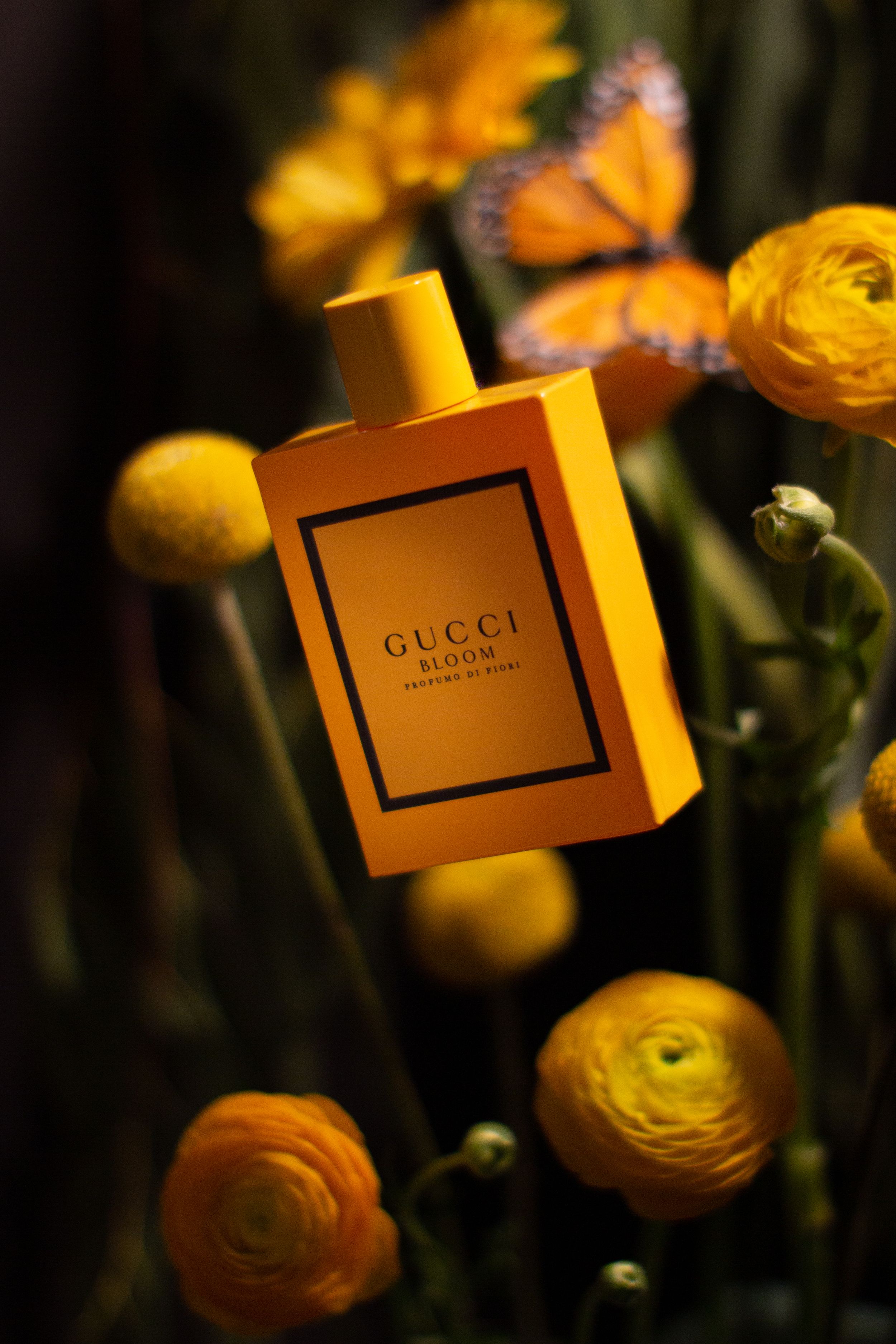Gucci perfume bottle surrounded by orange ranunculus flowers and a butterfly in warm lighting.