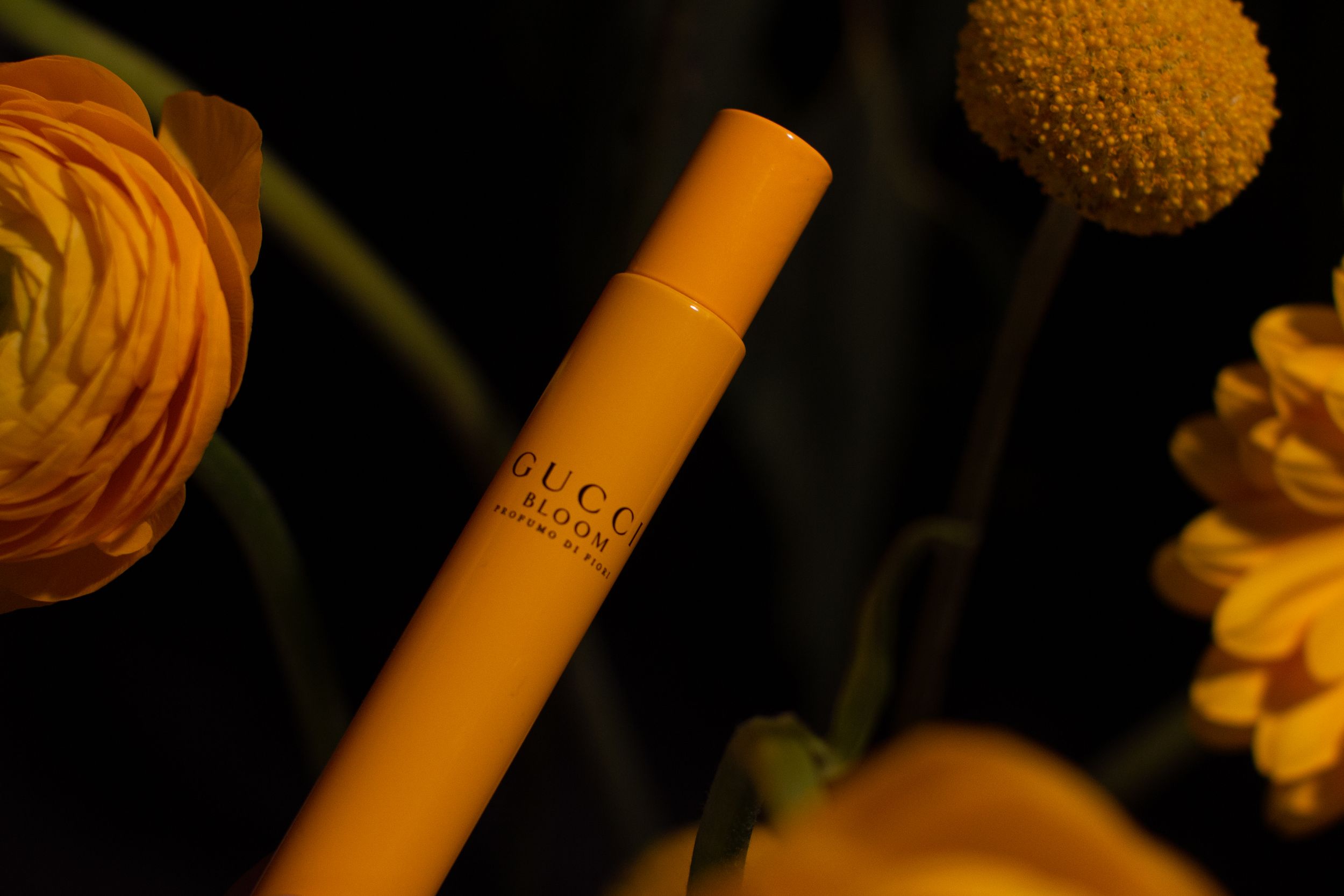 Close-up of an orange Gucci beauty product against a dark background with blurred flowers.