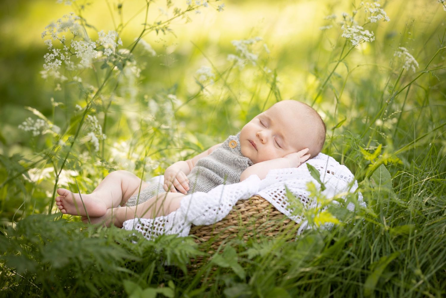 Baby sleeping peacefully in wicker basket among wildflowers and grass.