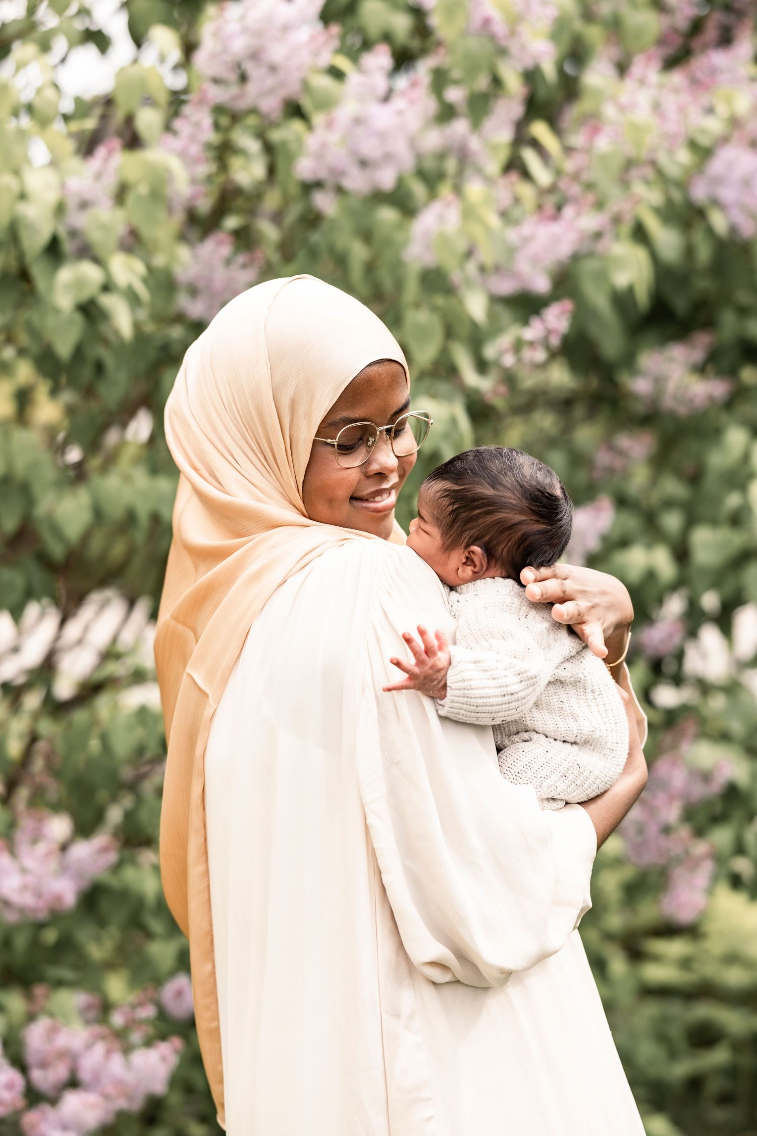 Woman in cream hijab holds and cherishes sleeping infant in garden with lilac blooms.