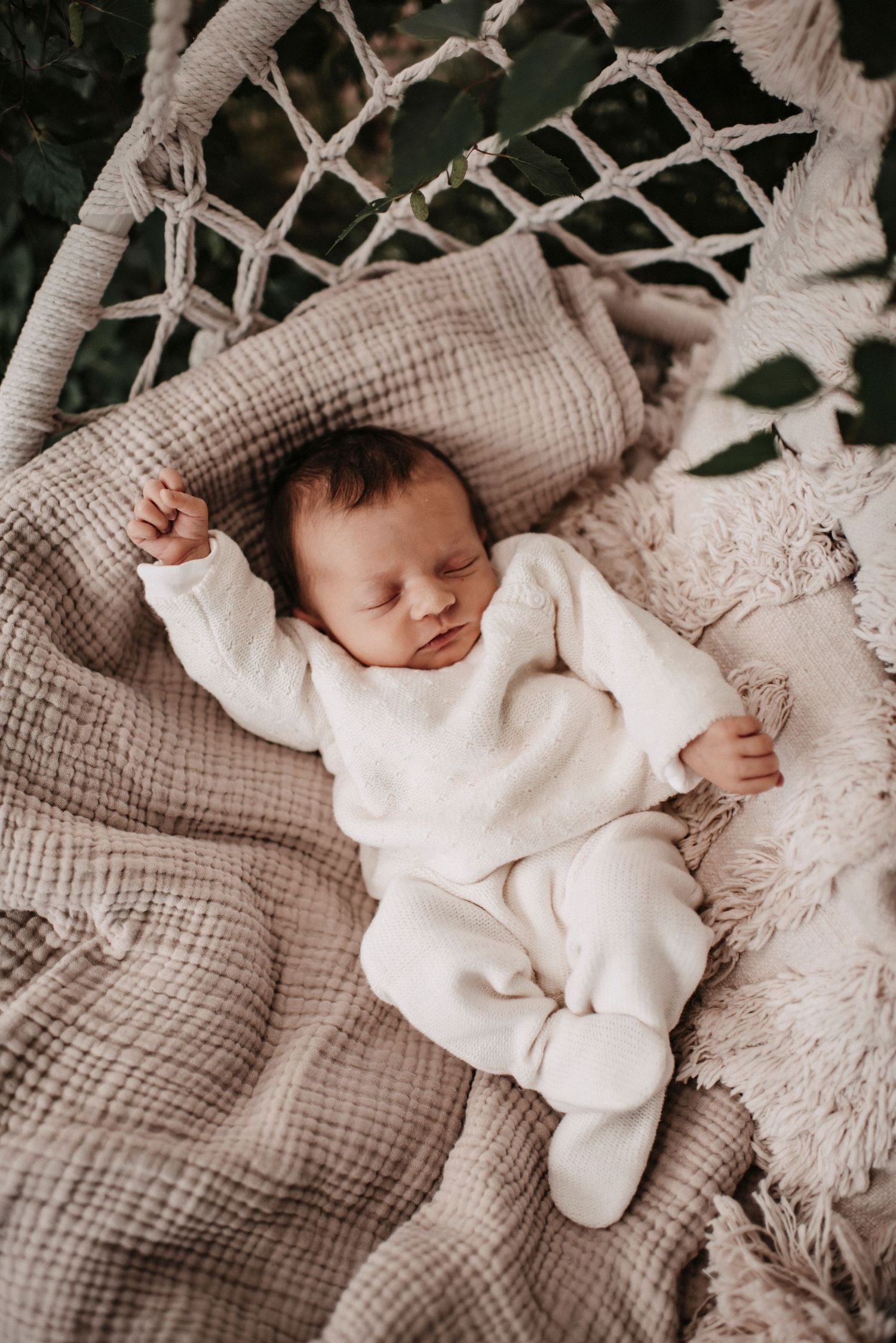 A peaceful baby in white sleeps on textured beige bedding in a macrame bassinet.