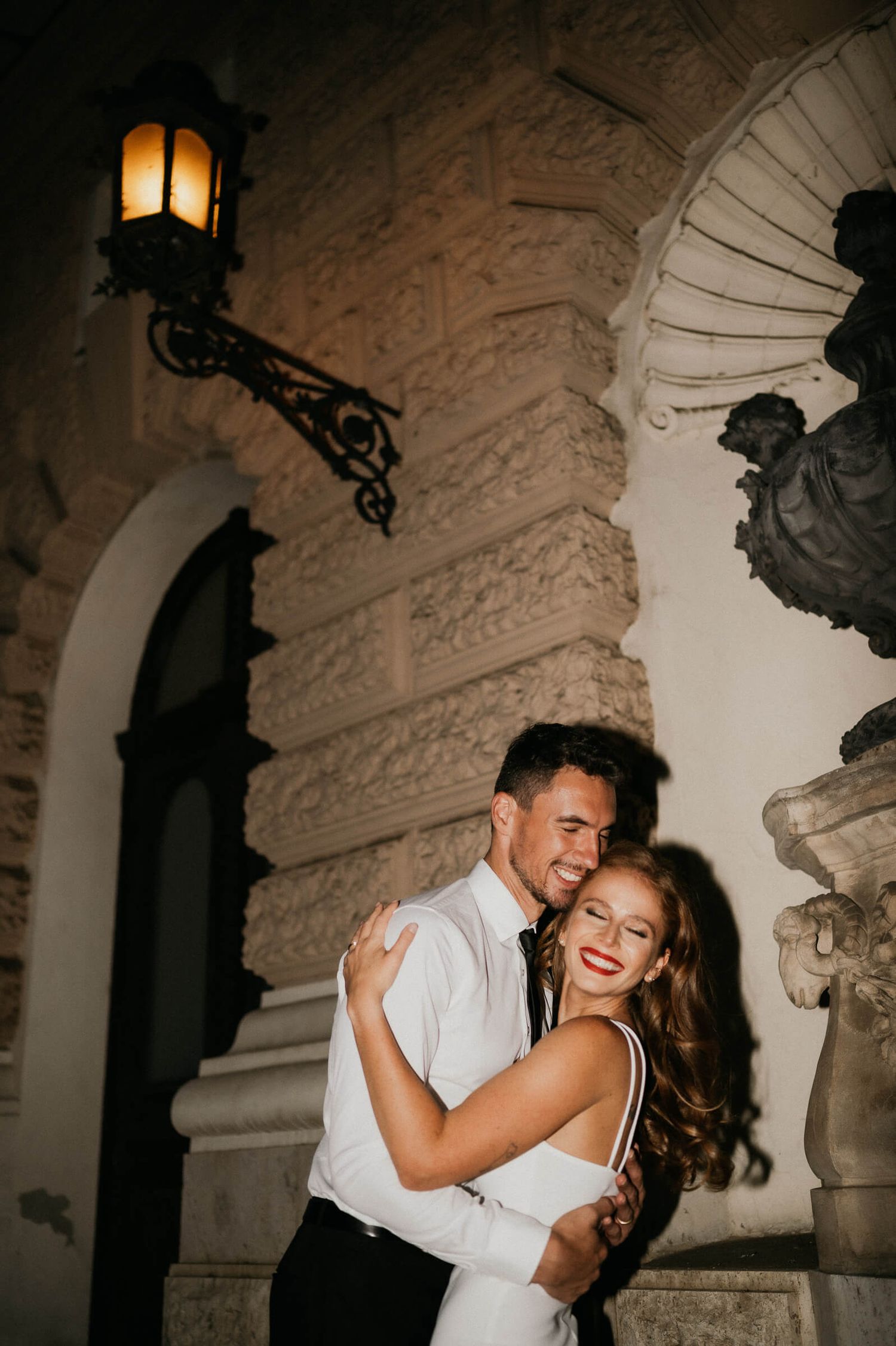 A romantic couple embrace and laugh together under an ornate wall sconce in an elegant architectural setting.