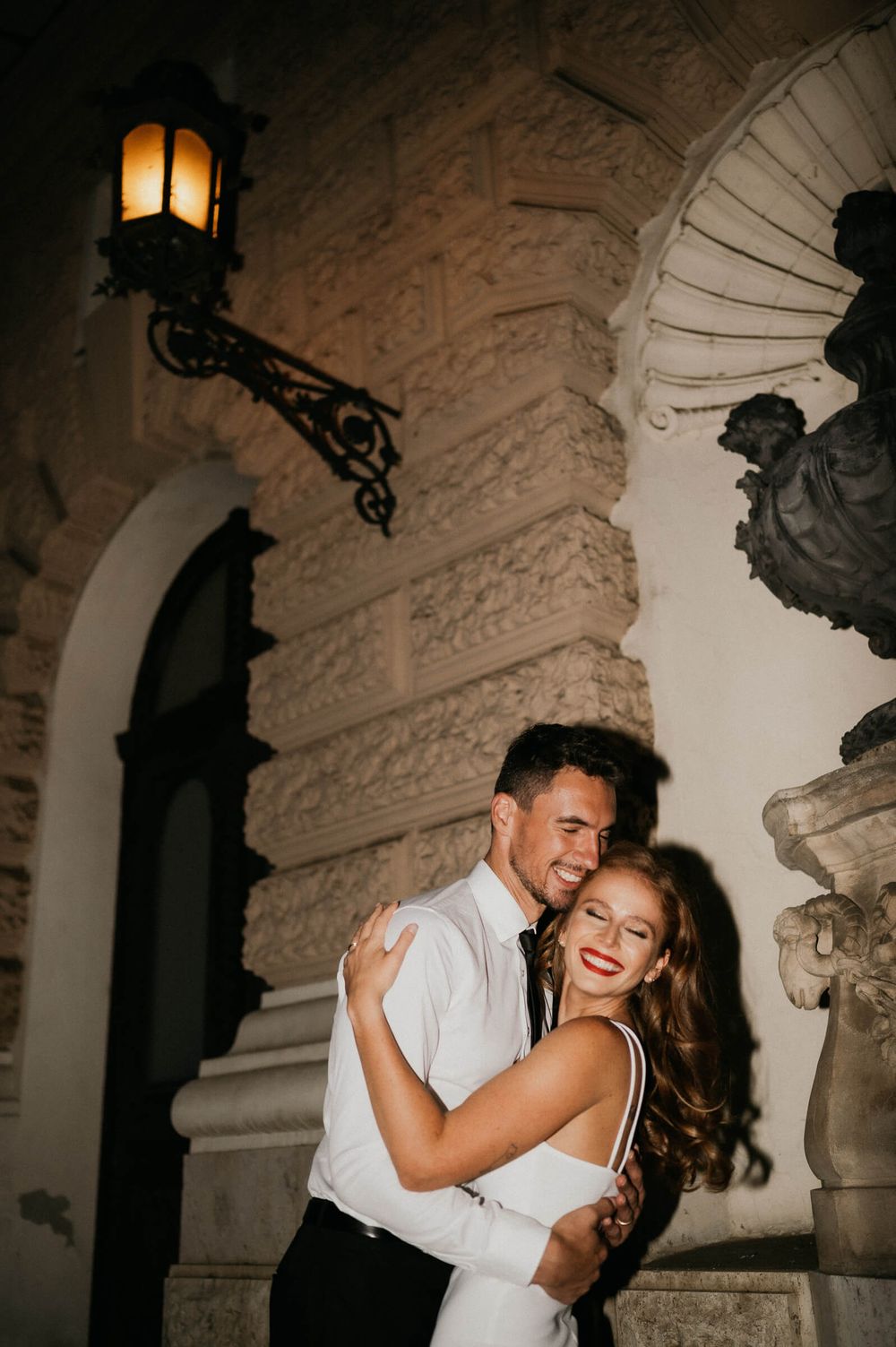 A romantic couple embrace and laugh together under an ornate wall sconce in an elegant architectural setting.