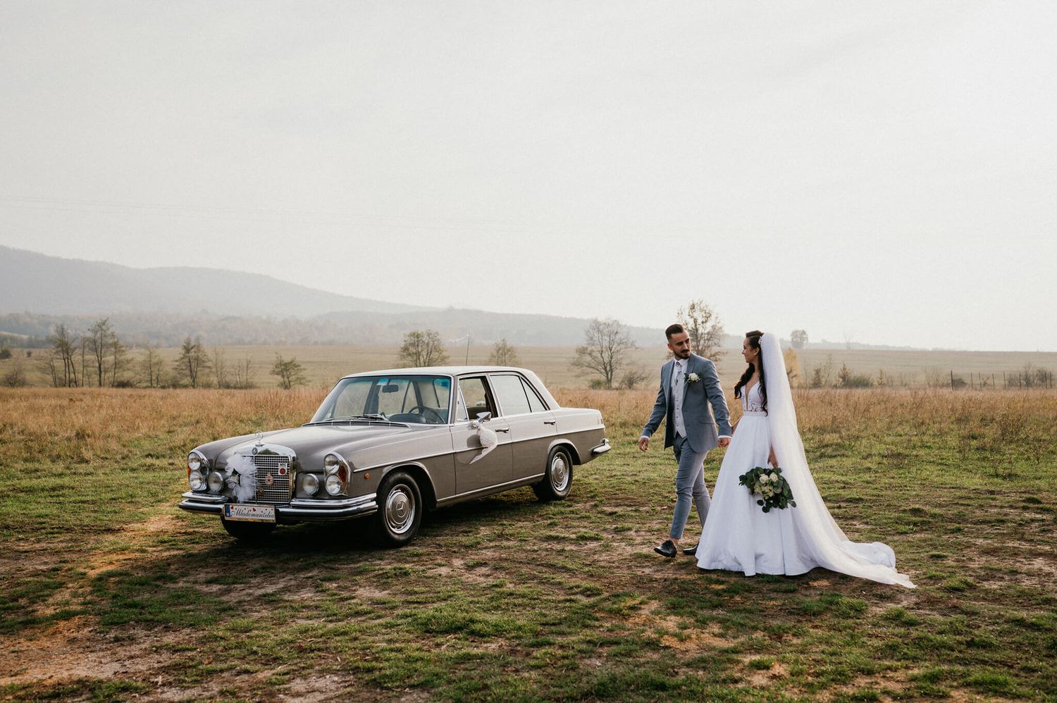 A classic Mercedes-Benz sits in a field as newlyweds pose beside it on their wedding day.