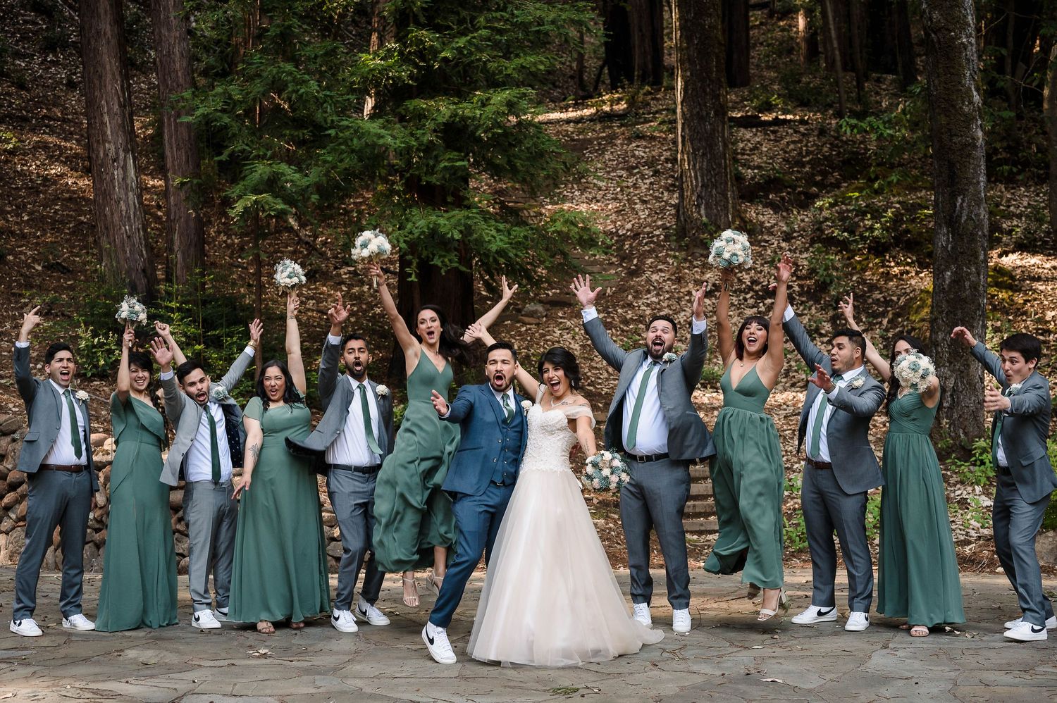 Wedding party in sage green dresses and suits celebrates joyfully in a forest setting.