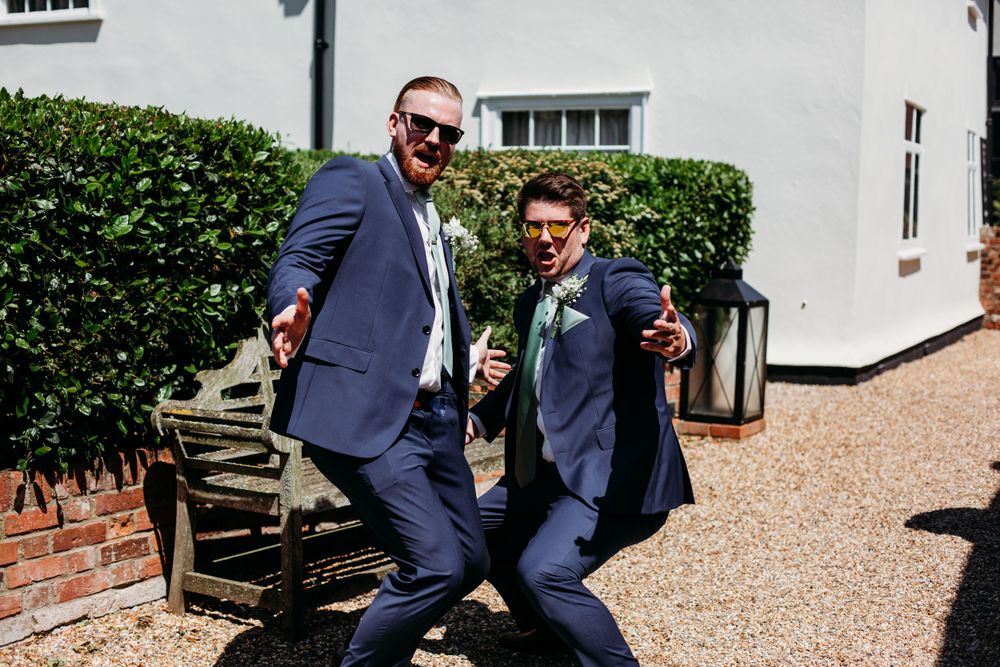Two men in navy suits striking fun poses on gravel driveway.