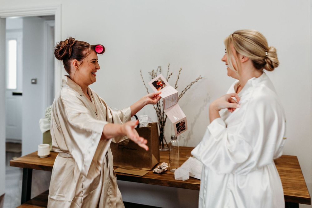 Two women in white bathrobes sharing drinks during wedding preparations.