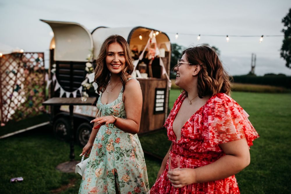 Two women in floral dresses share a laugh outdoors during a summer event.