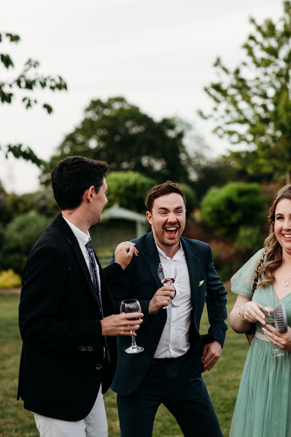 Candid moment of wedding guests laughing and enjoying drinks in garden setting.
