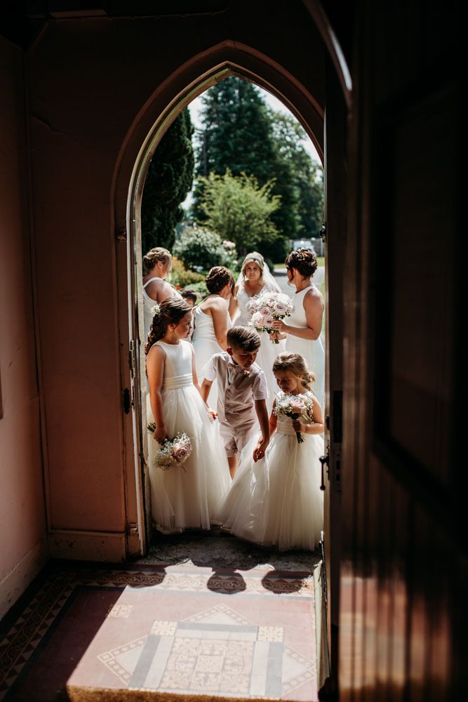 Bridesmaids gather in white dresses through an arched church doorway.