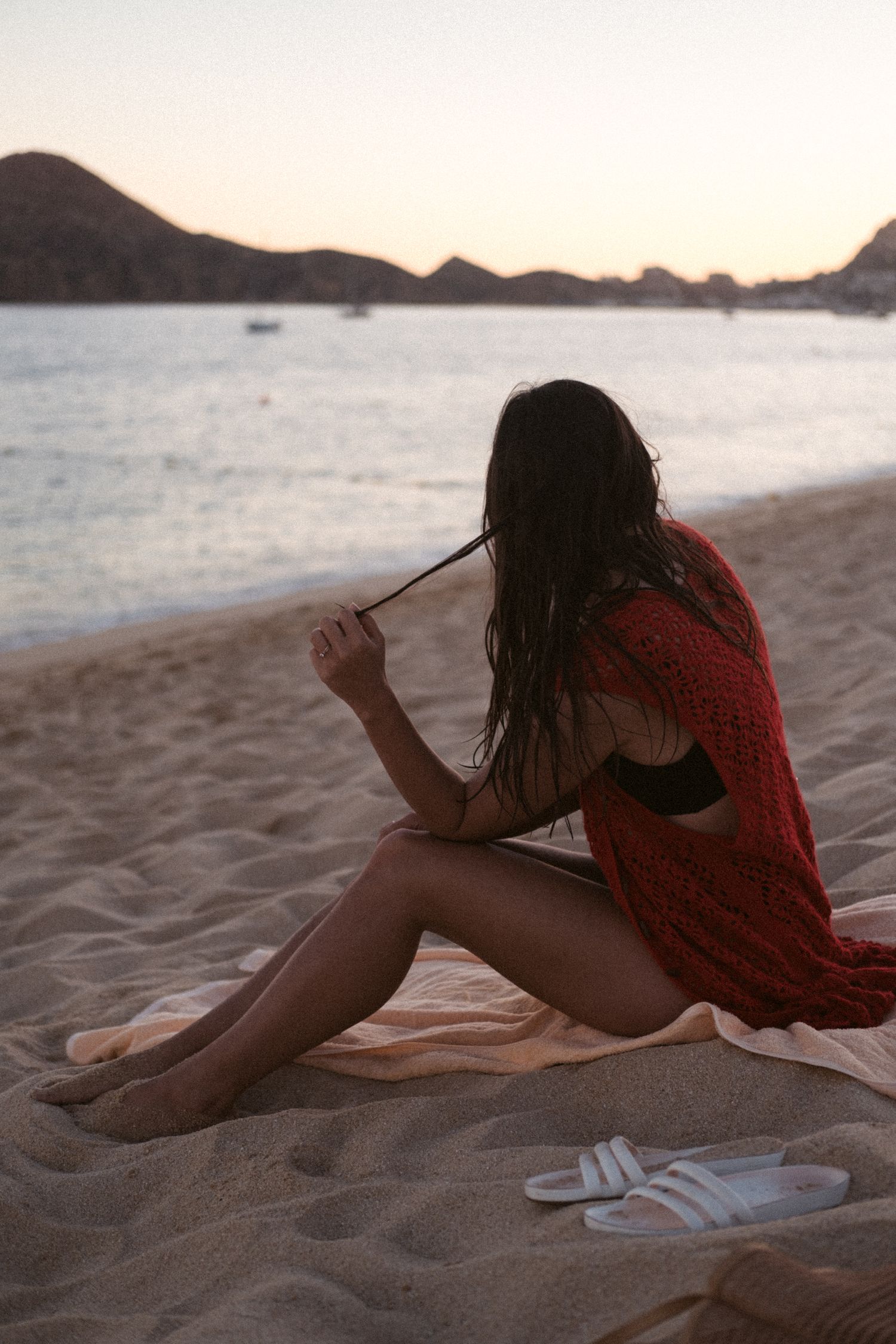 Person relaxing on sandy beach at sunset with sandals nearby.