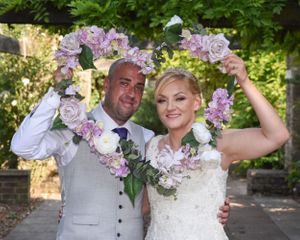 A joyful wedding couple poses outdoors with pink and purple floral arrangements.
