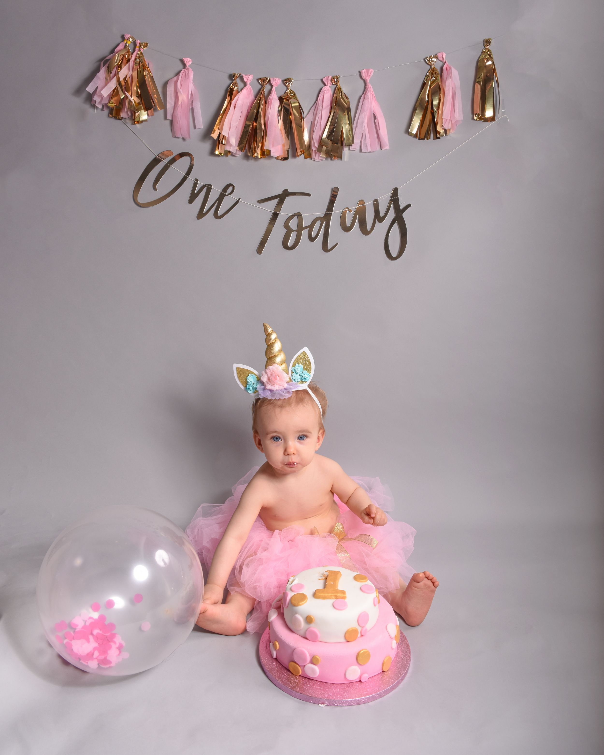 First birthday cake smash setup with pink decorations, balloons, and tassel garland against grey backdrop.