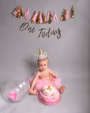 First birthday cake smash setup with pink decorations, balloons, and tassel garland against grey backdrop.