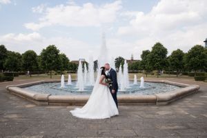 Wedding couple poses by outdoor fountain creating panoramic romantic scene.