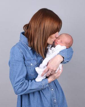 Person in denim shirt holding and kissing baby in white outfit against grey background.