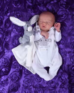 Newborn baby wearing white outfit lying on purple textured backdrop during professional photo shoot.