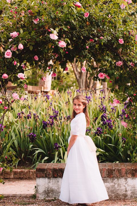 Full length portrait of girl in white dress standing among roses and purple iris flowers.