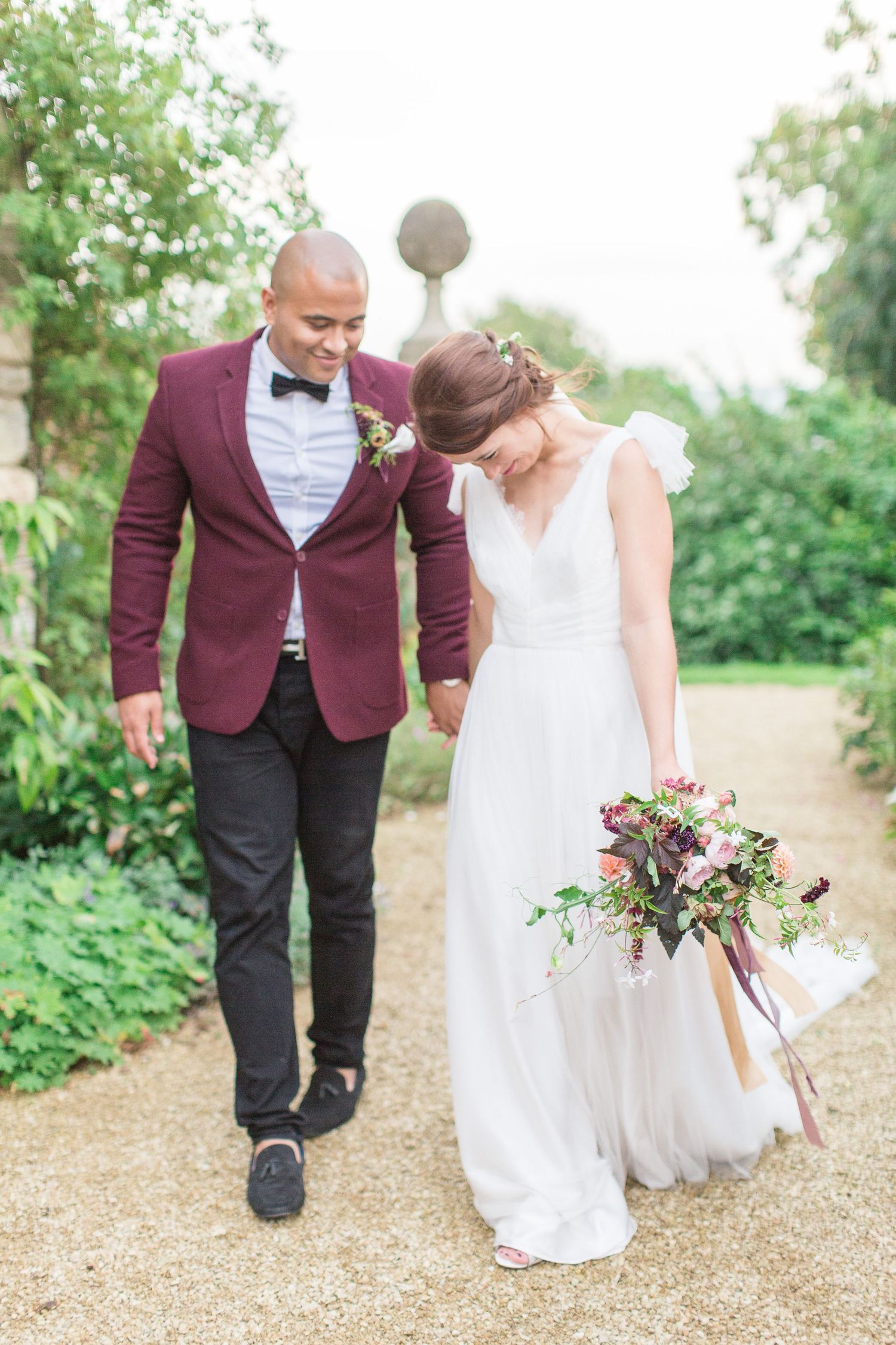 A couple in wedding attire walks along a garden path, with the bride holding a pink and green floral bouquet euridge manor