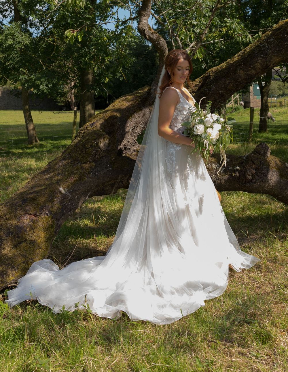 Bride in flowing white dress poses with bouquet against rustic tree in sunlit garden.
